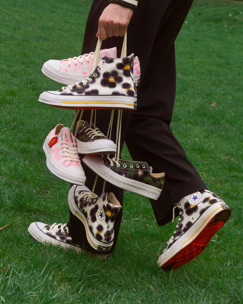 A person carries multiple pairs of Converse shoes in a grassy outdoor setting, showcasing different styles and patterns.