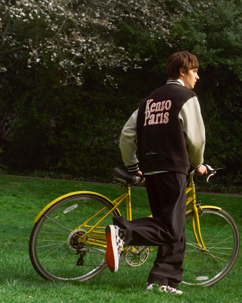 A young adult male stands with a yellow bicycle on green grass, wearing a black and white jacket and black pants in an outdoor setting.