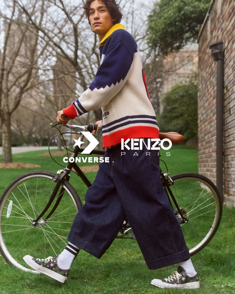 A young adult man in a sweater walks alongside a bicycle in an outdoor setting, branded with visible Converse and Kenzo logos.