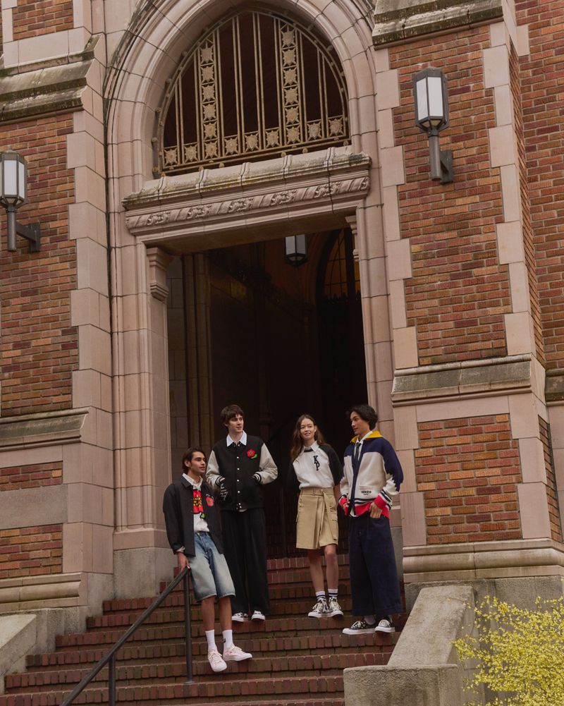 Four young adults in varsity jackets stand on brick steps outside a stone building with arched entrance in an urban setting. 