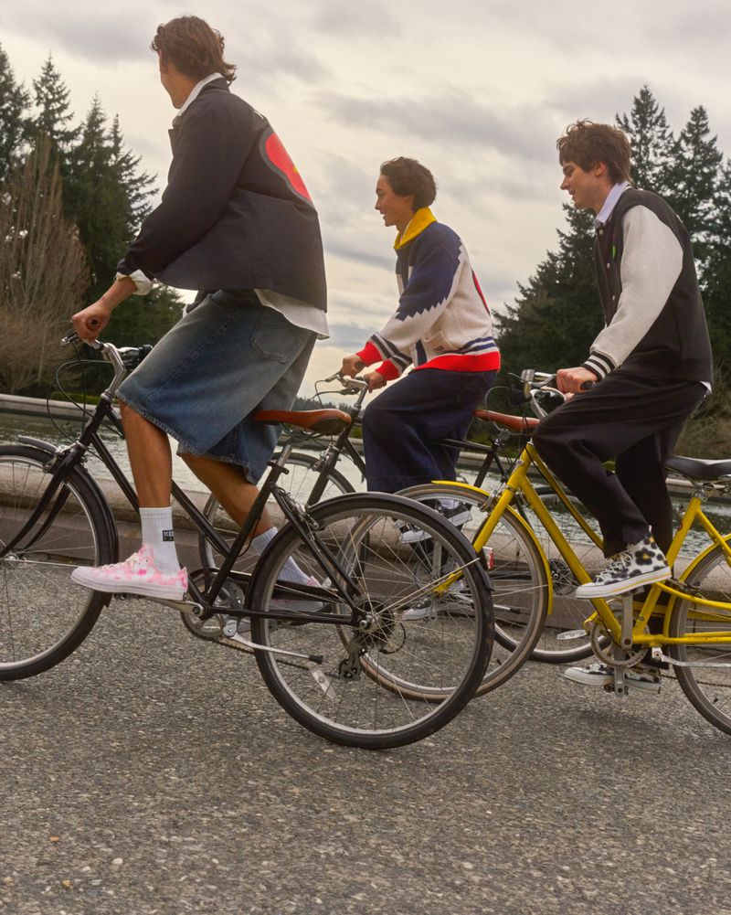 Three young adults ride bicycles on a paved path near a river; overcast sky, green trees, and casual clothing add to the outdoor scene.