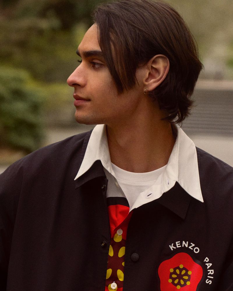 Profile of a young adult man with dark hair wearing a black jacket with a red and yellow floral design in an outdoor setting.