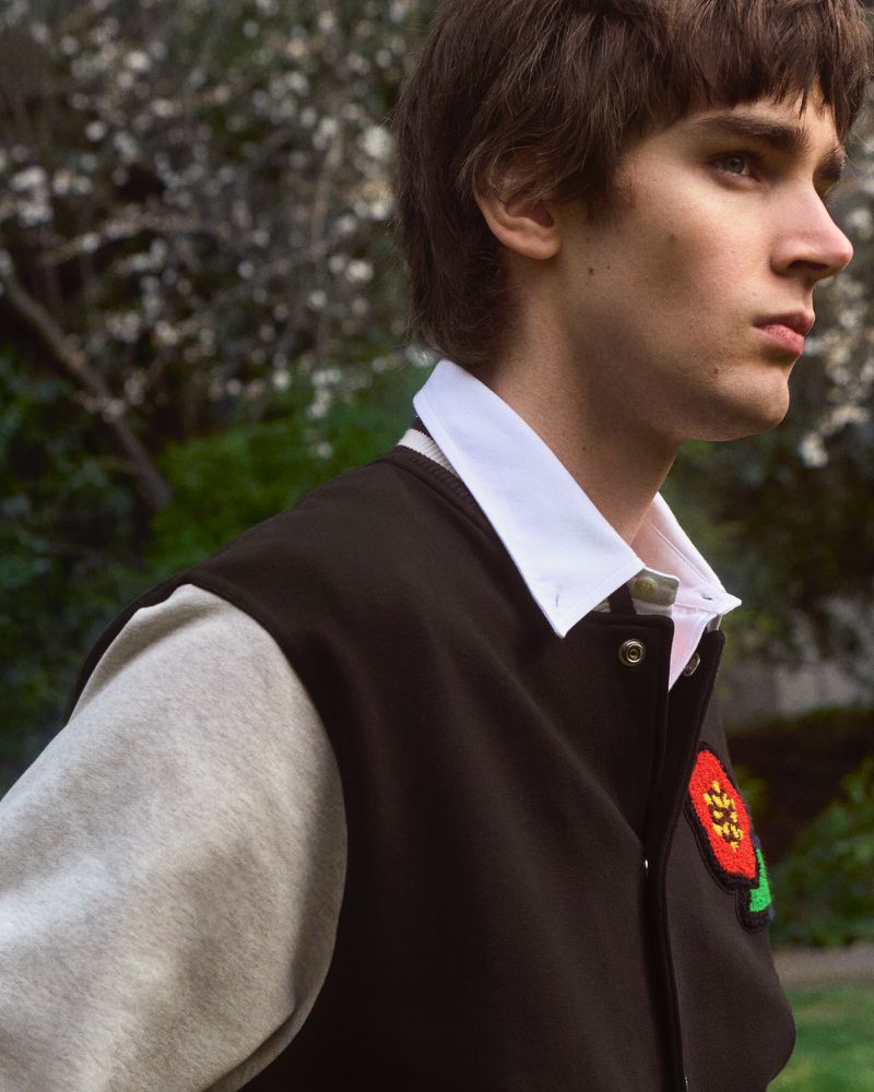 Profile of a young adult male with dark hair, wearing a varsity jacket, posing outdoors against a green foliage backdrop.