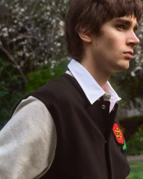 Young male model in a dark varsity jacket with a red flower patch over a white collared shirt, photographed outdoors.