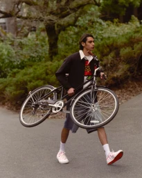 Model carrying a bicycle while walking along a paved park path in the Kenzo x Converse Campaign