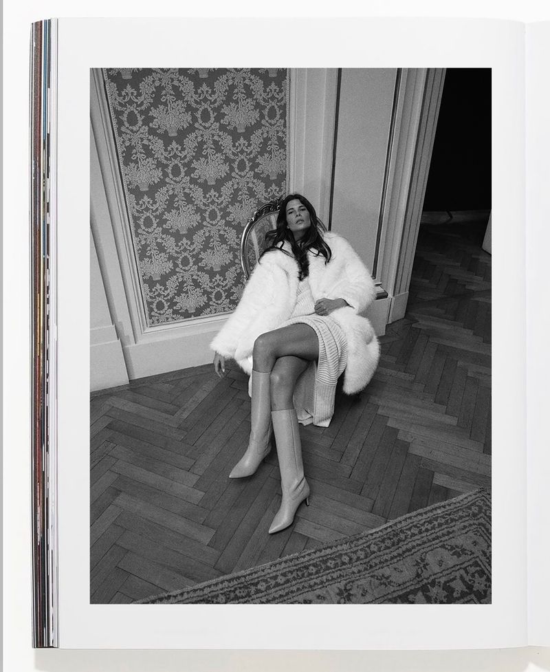 Black and white magazine photo of a woman in fur coat and boots sitting on a chair in a room with herringbone floor.