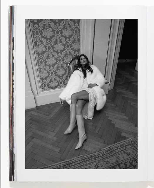 Black and white magazine photo of a woman in fur coat and boots sitting on a chair in a room with herringbone floor.