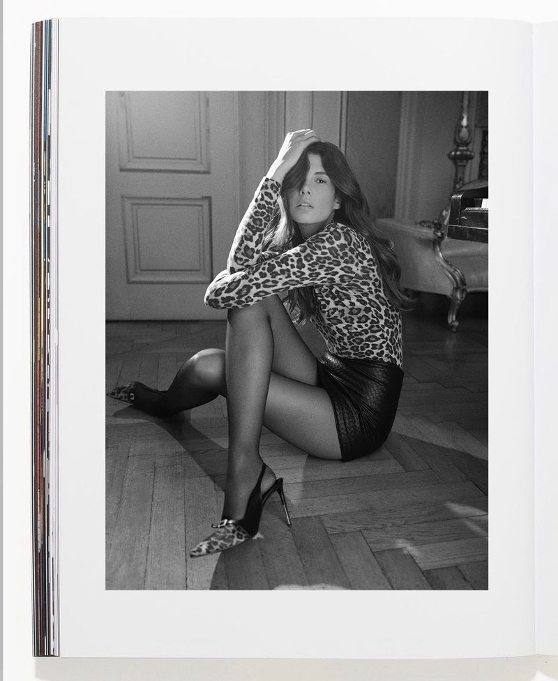 A woman in a leopard print shirt and black mini skirt sits on the floor in a black and white photograph, posed with hands in hair.