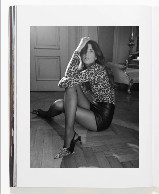 A woman in a leopard print shirt and black mini skirt sits on the floor in a black and white photograph, posed with hands in hair.