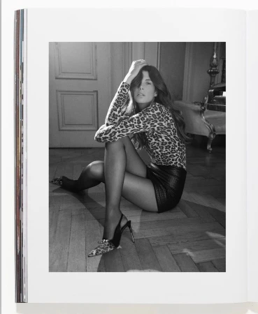 A woman in a leopard print shirt and black mini skirt sits on the floor in a black and white photograph, posed with hands in hair.