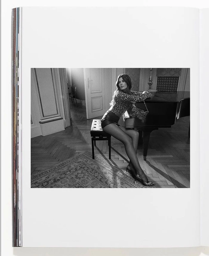 Black and white editorial portrait of Monica Cima seated on a piano bench next to a grand piano in an elegant room.
