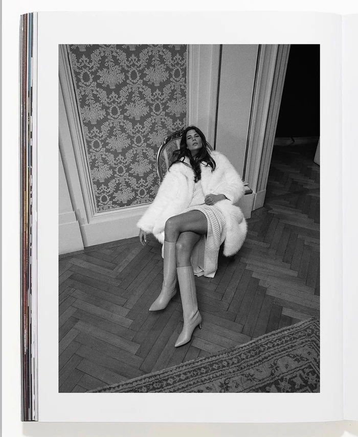 Model wearing a white faux fur coat and tall boots seated on an ornate chair in a room with patterned wallpaper and parquet flooring.