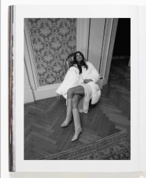 Model wearing a white faux fur coat and tall boots seated on an ornate chair in a room with patterned wallpaper and parquet flooring.