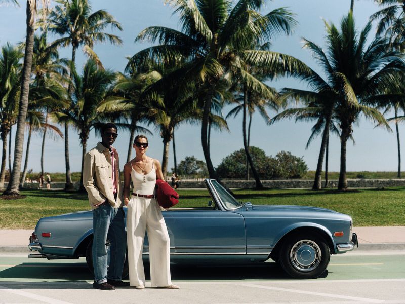 A fashionable couple poses next to a vintage convertible car on a sunny day with palm trees in the background, evoking a sense of retro style.