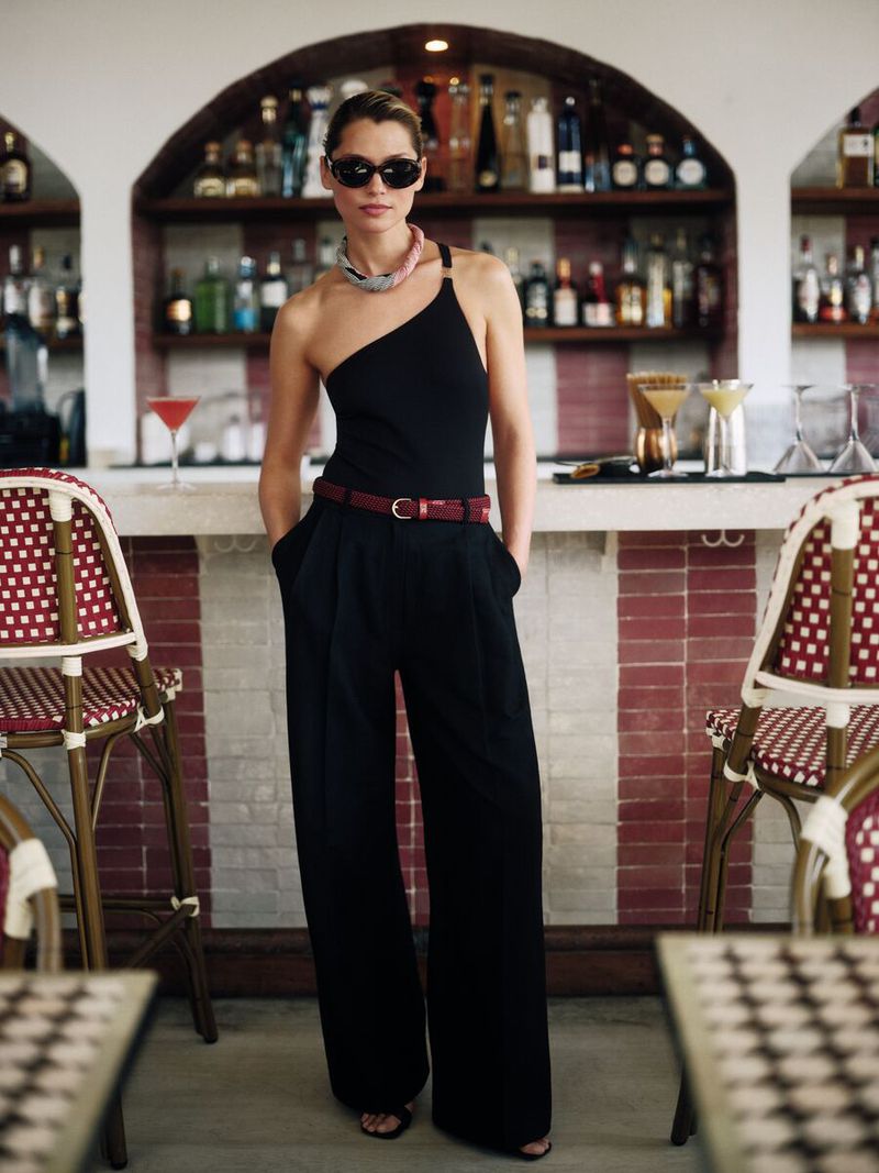 Elegant woman in black one-shoulder jumpsuit with burgundy belt poses in a bar setting with red and white striped accents.