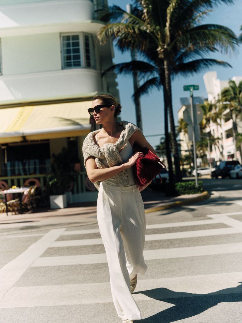 A woman in white pants and a sweater walks across a zebra-striped crosswalk, holding a burgundy purse, with palm trees in the background.