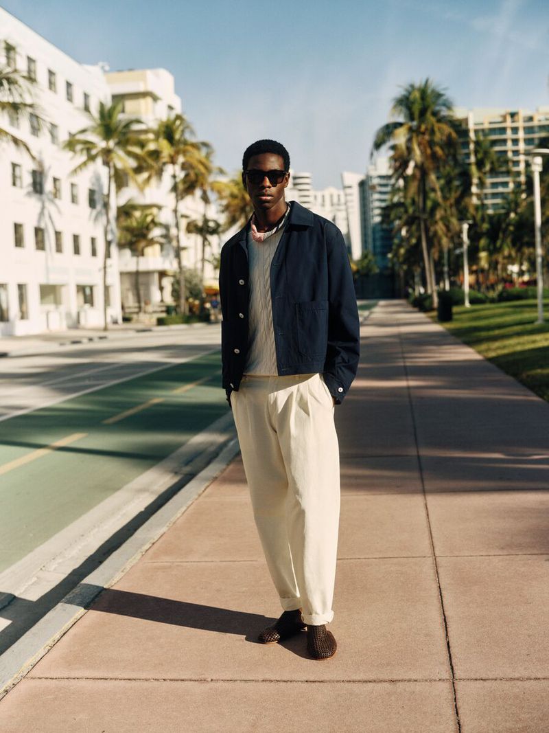 Man wearing a dark jacket and light trousers standing on a palm-lined sidewalk under clear daylight
