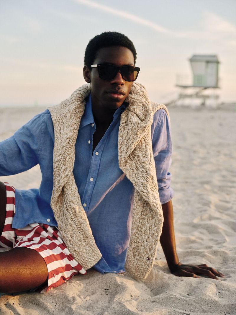 A young man in a light blue shirt and red and white striped shorts sits on a sandy beach with a lifeguard tower in the distance.