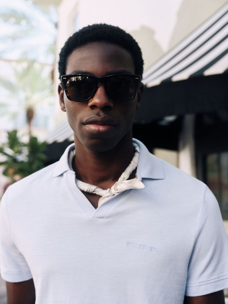 Portrait of a young Black man wearing sunglasses and a light blue polo shirt outdoors with green palm trees in the background.