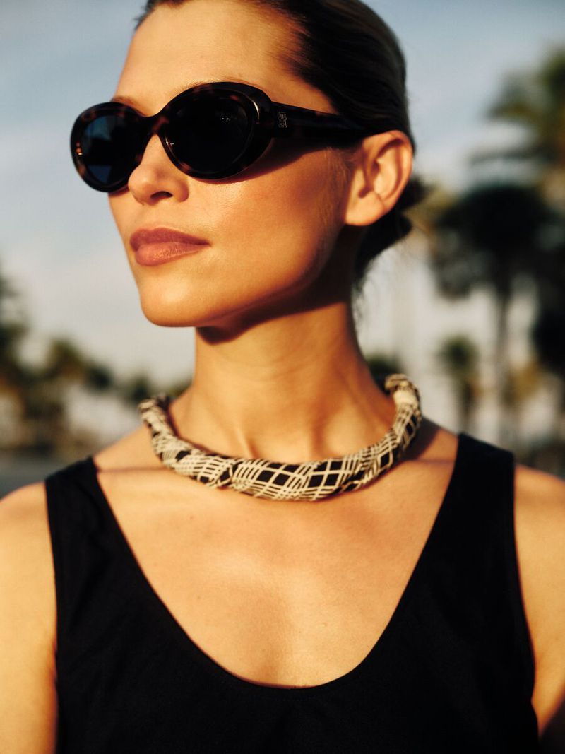 A fashionable adult woman with sunglasses and a necklace poses in front of palm trees, bathed in warm, golden sunlight.