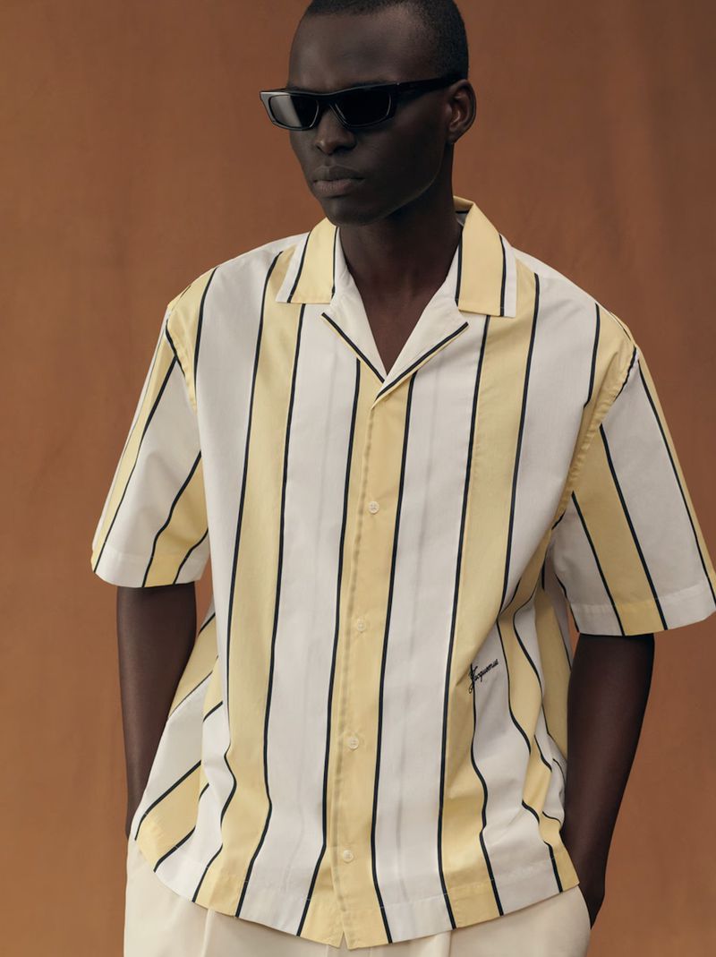 Dark-skinned male model poses in an unbuttoned white and yellow striped shirt, paired with off-white pants against a neutral background.