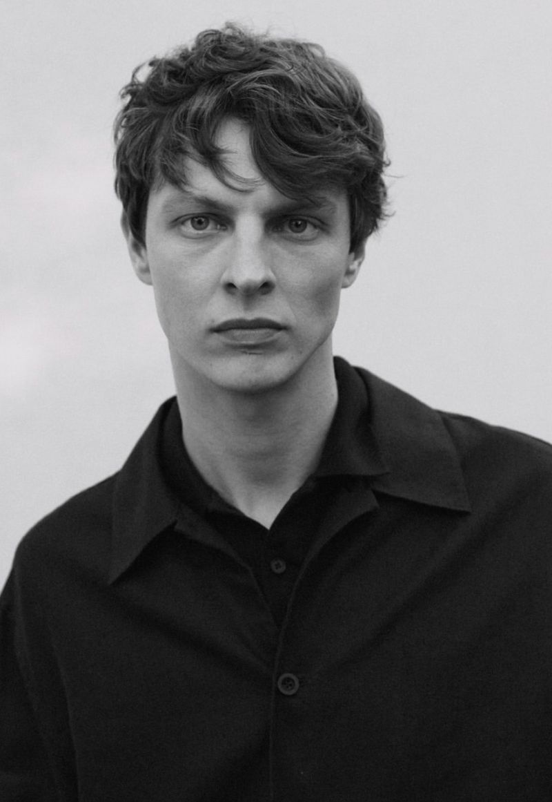 Black-and-white portrait of a young adult male with curly hair, wearing a dark collared shirt, against a bright, neutral background.