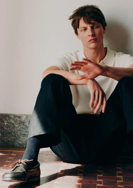 Man in polo and slacks seated indoors. Male fashion shot with dark hair and dark socks, wearing casual shoes in warm indoor light.