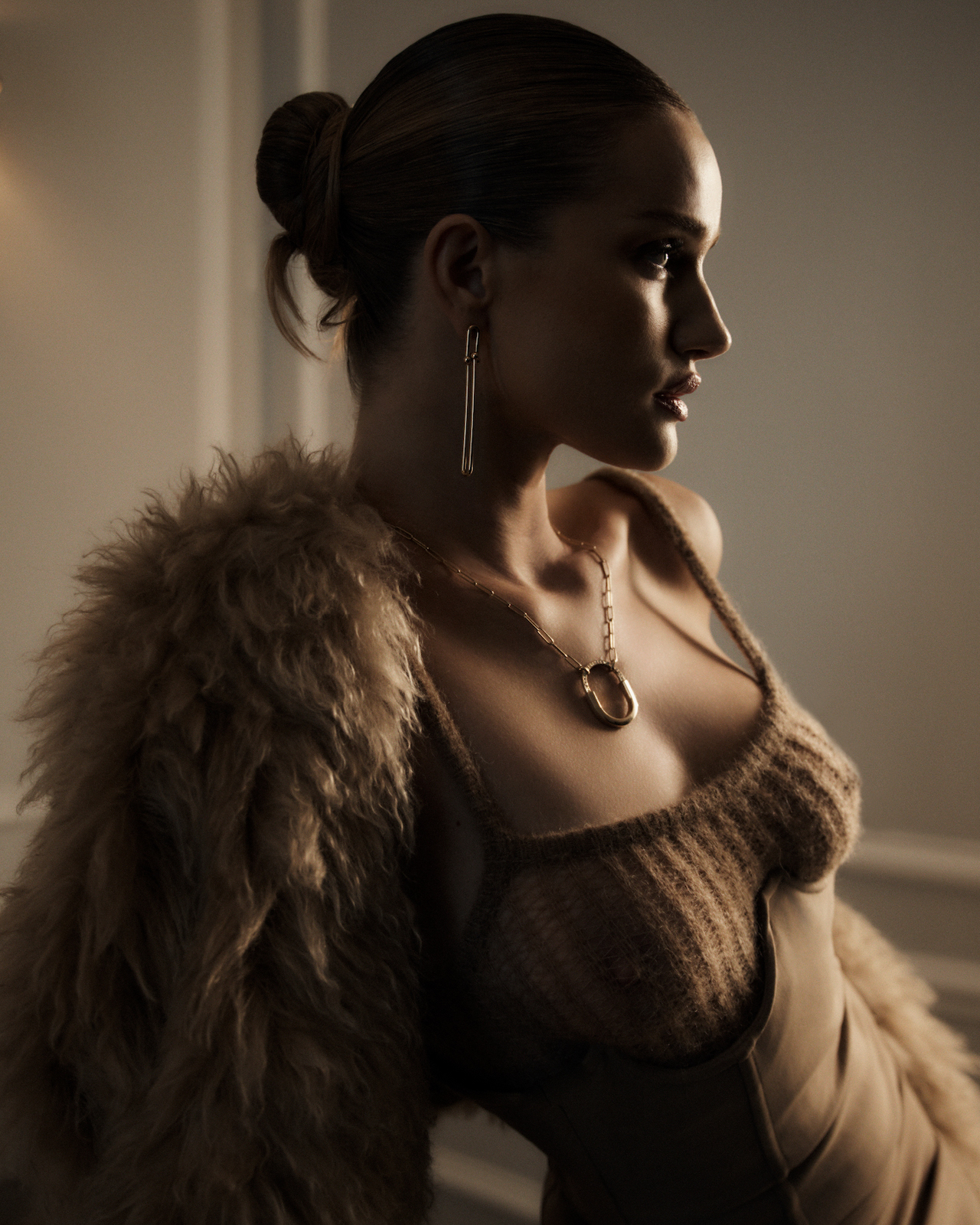 Portrait of a woman in a knit top and fur coat looking away, featuring neutral tones, gold jewelry, and an indoor environment with soft lighting.