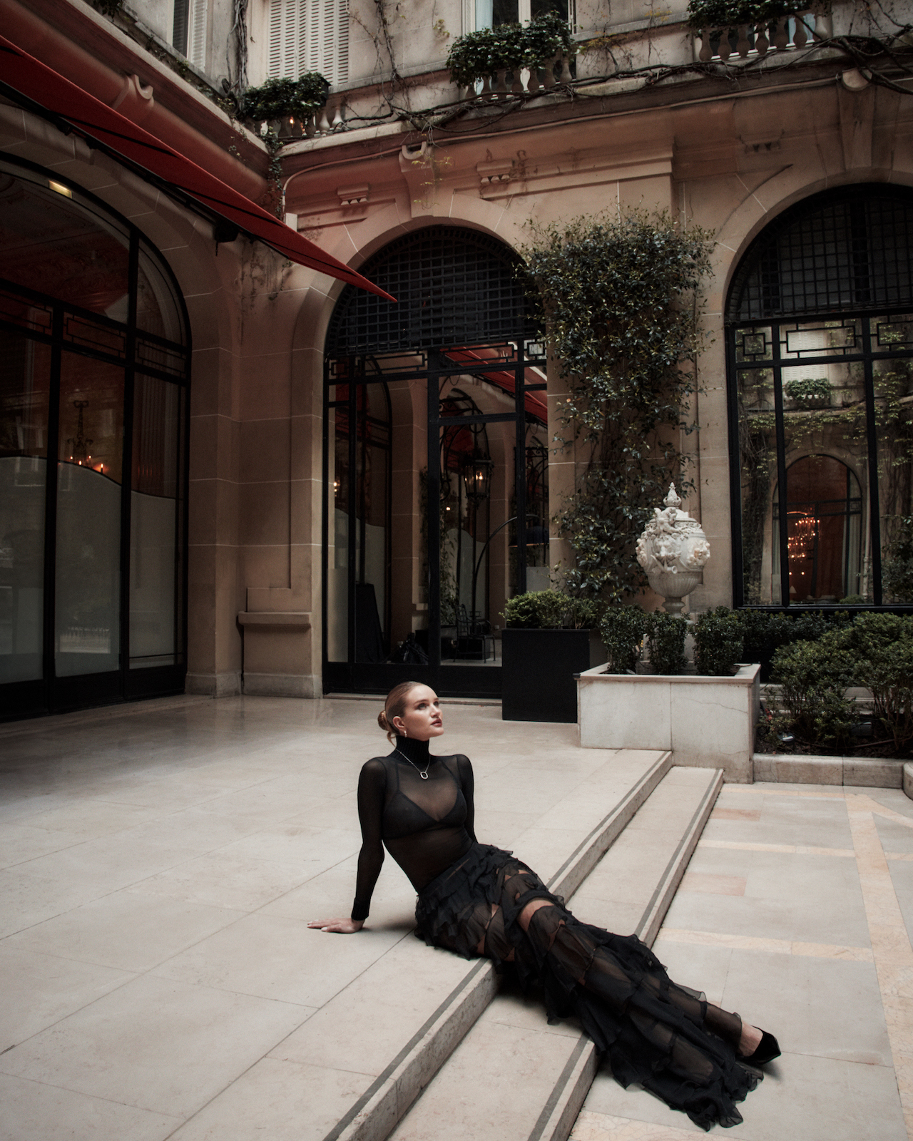 Fashion shot of an adult woman in a black dress, seated on stone stairs in a courtyard of a stone building with arched windows.