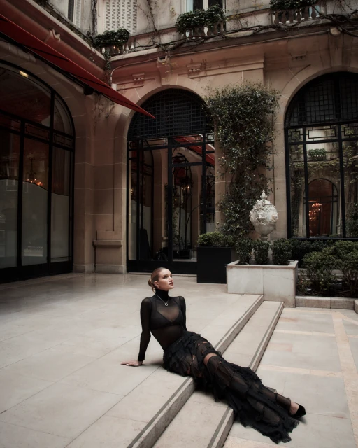 Fashion shot of an adult woman in a black dress, seated on stone stairs in a courtyard of a stone building with arched windows.