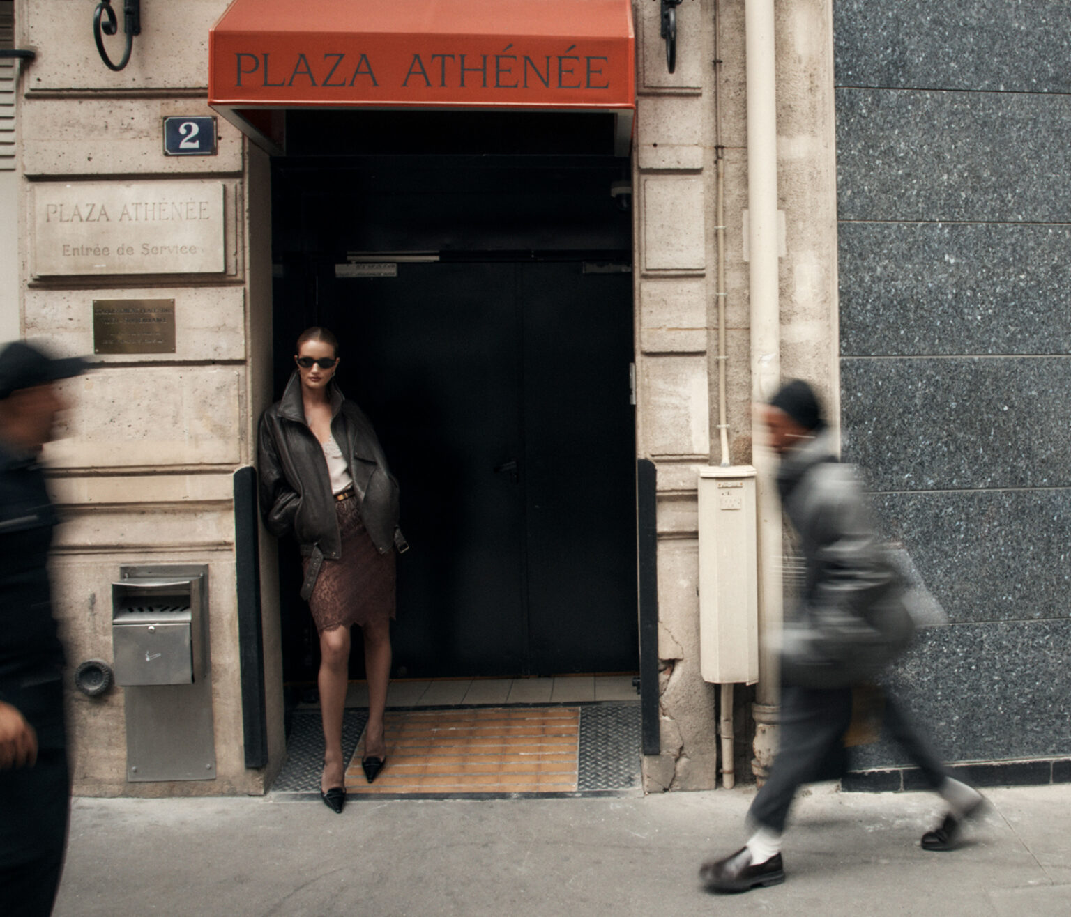 Woman in dark sunglasses and leather jacket leaning in a doorway beneath a Plaza Athenee sign with a blurred figure walking past