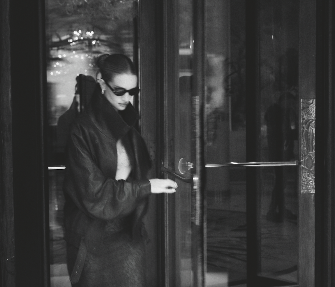 Monochrome portrait of a woman opening a glass door, wearing sunglasses, a leather jacket, and skirt in an indoor setting.
