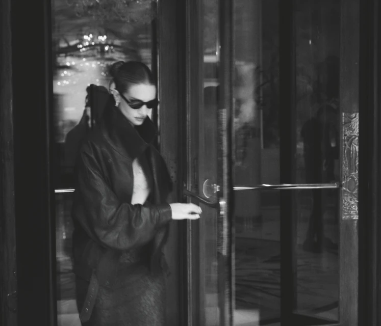 Monochrome portrait of a woman opening a glass door, wearing sunglasses, a leather jacket, and skirt in an indoor setting.