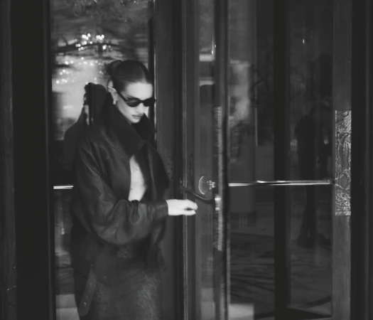 Monochrome portrait of a woman opening a glass door, wearing sunglasses, a leather jacket, and skirt in an indoor setting.