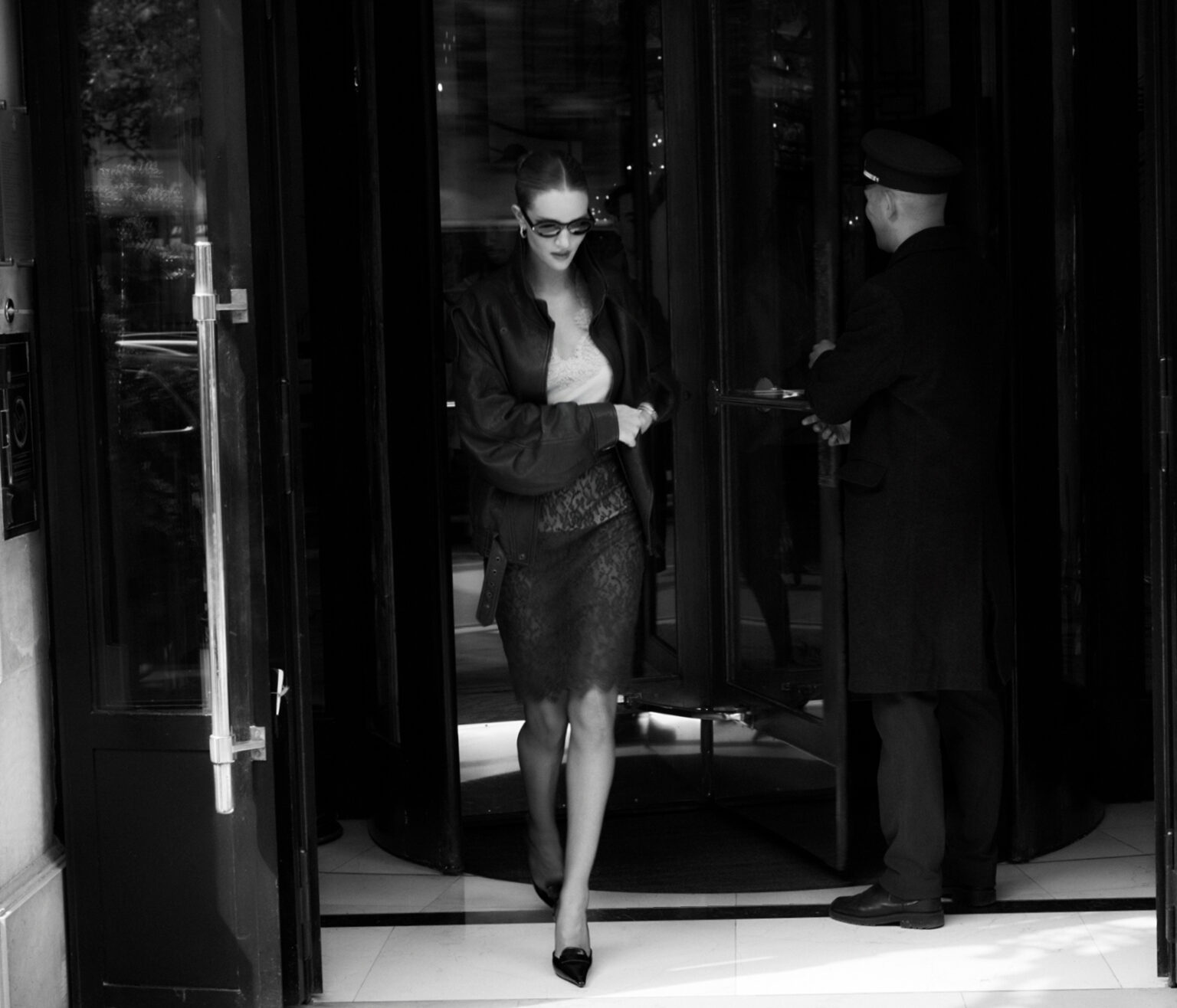 Elegant woman in a dark jacket and skirt exiting building, greeted by doorman, black and white photography, eye-level angle.