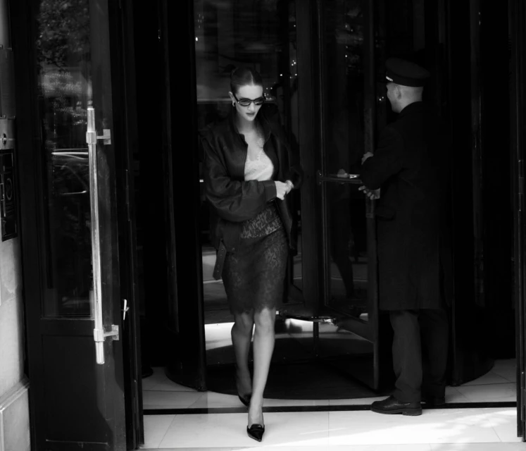 Elegant woman in a dark jacket and skirt exiting building, greeted by doorman, black and white photography, eye-level angle.