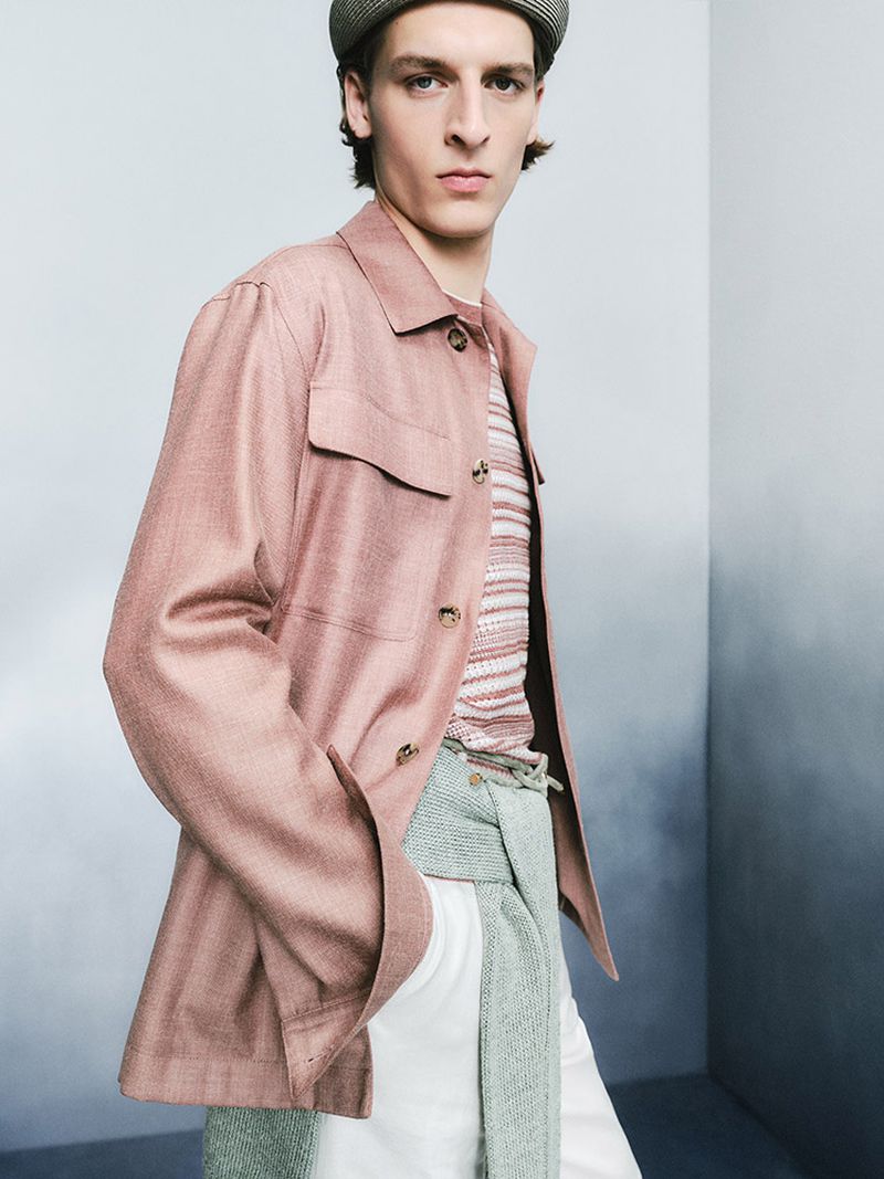 Portrait of a male fashion model wearing a rose-colored jacket, striped shirt, cap, and white pants against a gradient backdrop.