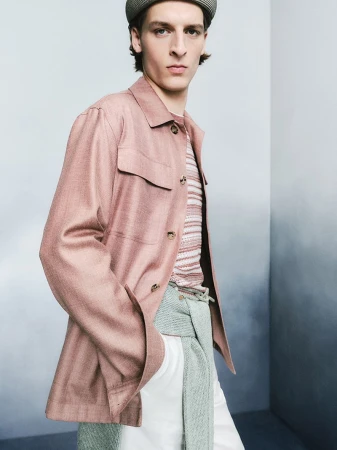 Male model wearing a pink jacket, striped shirt, and flat cap against a soft gradient studio backdrop.