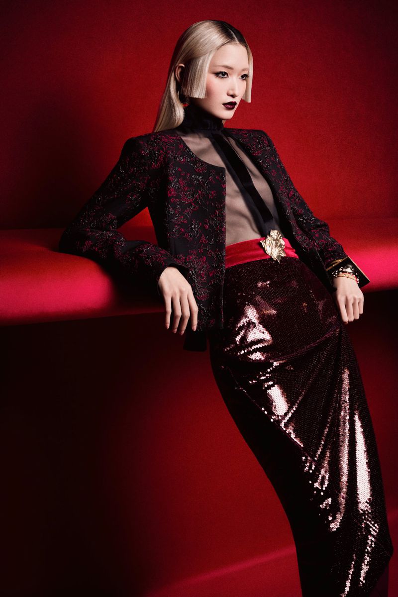 Elegant Asian woman in a dark floral jacket and sequin skirt poses against a red backdrop, showcasing a high-fashion look with dramatic makeup.