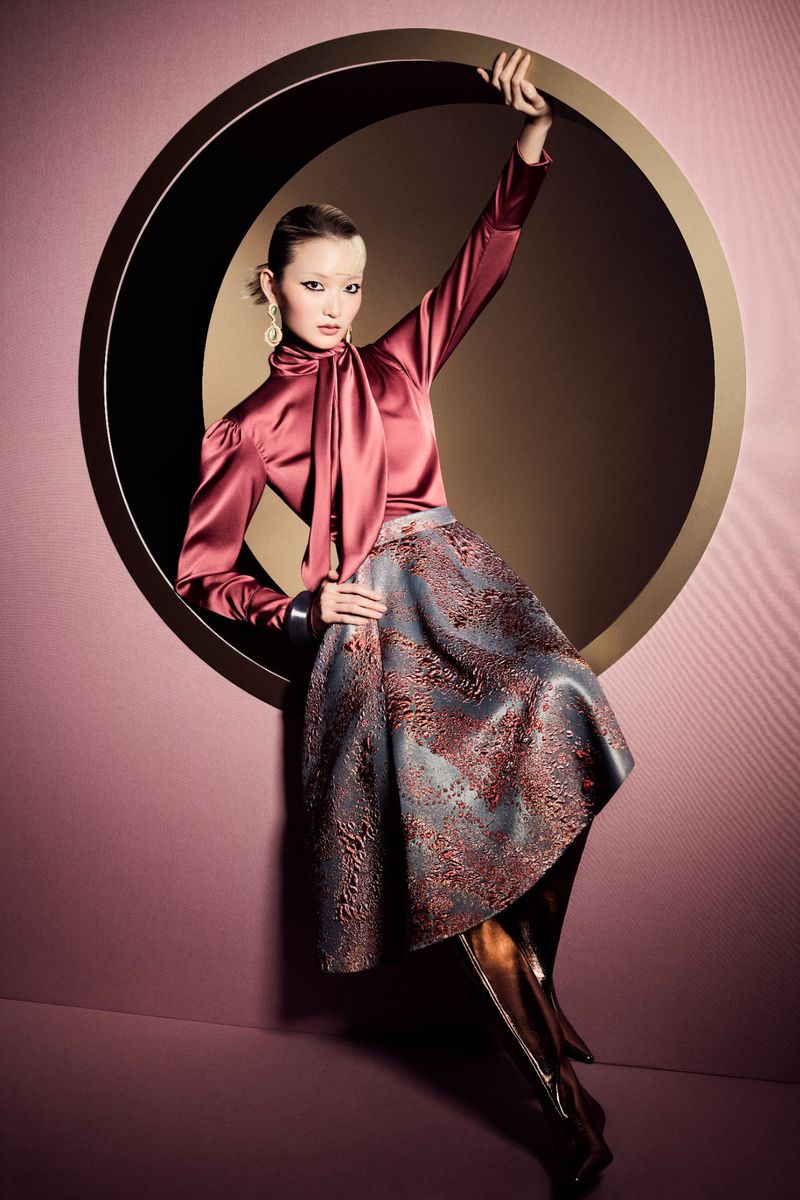 Elegant woman poses inside a circular window wearing a satin blouse, patterned skirt, gold earrings, and metallic knee-high boots in a studio.