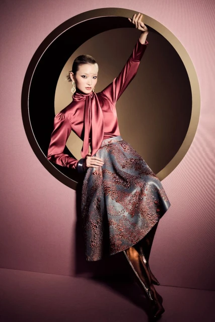 Elegant woman poses inside a circular window wearing a satin blouse, patterned skirt, gold earrings, and metallic knee-high boots in a studio.