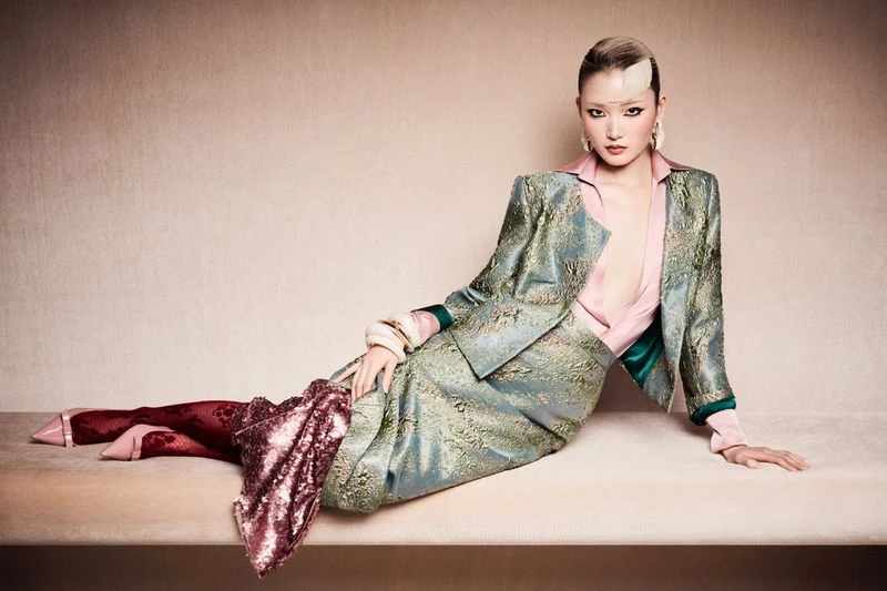 A stylish Asian woman in a teal suit poses on a beige bench, illuminated by soft lighting against a neutral backdrop, fashion shoot.