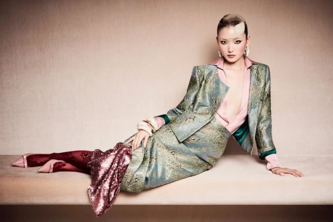 A stylish Asian woman in a teal suit poses on a beige bench, illuminated by soft lighting against a neutral backdrop, fashion shoot.