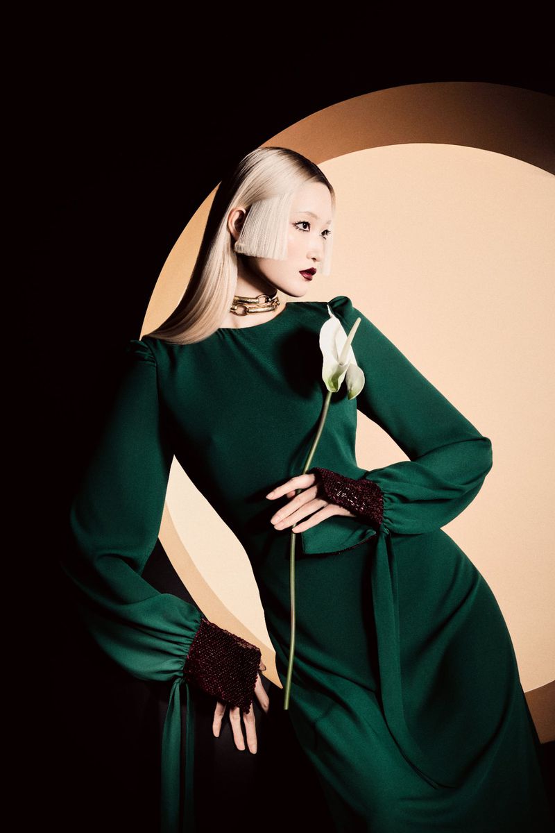 Elegant Asian woman in a green gown poses against a geometric background, holding a white calla lily in a fashion photography shot.