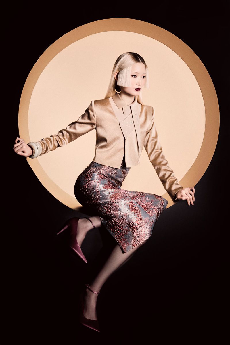 Elegant Asian woman posing in a studio. She wears a stylish jacket, skirt, and heels, set against a striking black and gold backdrop.