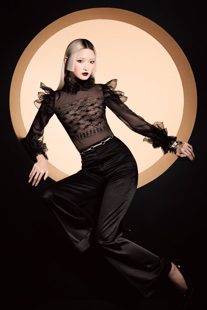 Stylish woman with platinum blonde hair posing in a sheer black blouse, black pants and black heels against a peach and black background.