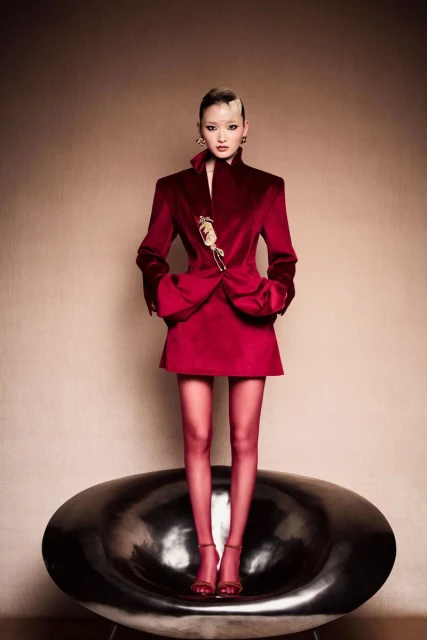 Full-length portrait of an Asian woman wearing a red velvet suit and standing on a modern black chair in a studio setting.
