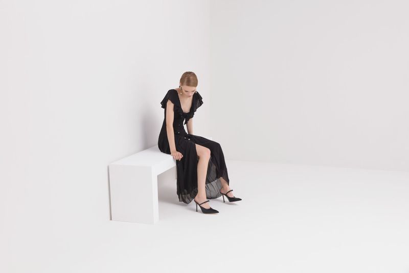 Blond woman sits on a white bench in a photo studio wearing a black dress and heels; the background is white, minimalist fashion photography.