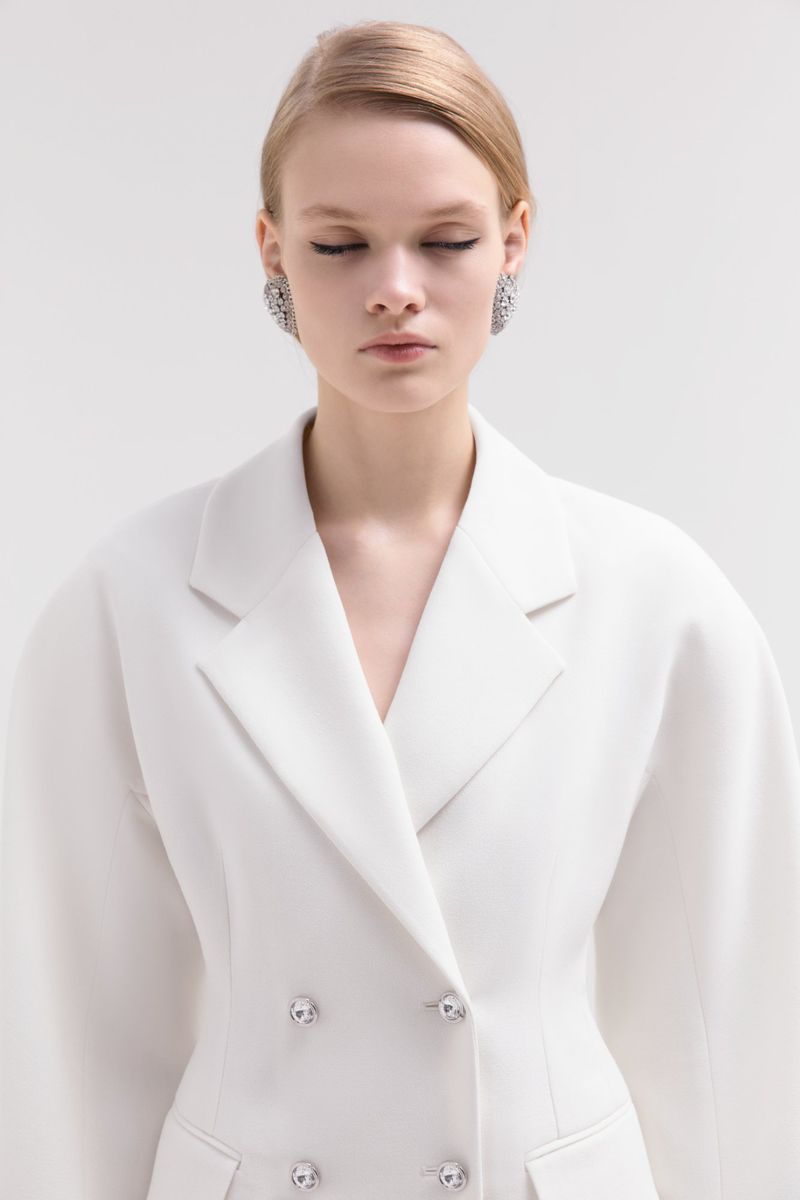 Woman in white double-breasted blazer with eyes closed. Studio shot of blond-haired, light-skinned model wearing statement earrings.