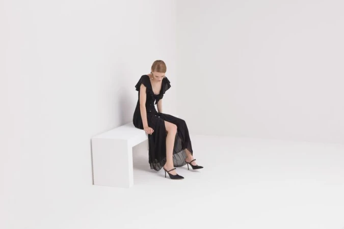 Model seated on a white block wearing a black ruffled dress and heels against a white backdrop